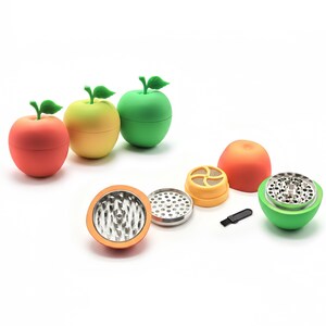 May include: A selection of colourful apple-shaped herb grinders in red, yellow, and green. The grinders are shown disassembled, revealing the internal components, including a metal grinding surface and a small black brush.