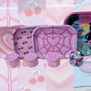 May include: A collection of four rectangular metal rolling trays and four matching cylindrical grinders. The designs include pink heart patterns, red cherry prints, purple spider web motifs with hearts, and a colorful graphic illustration.