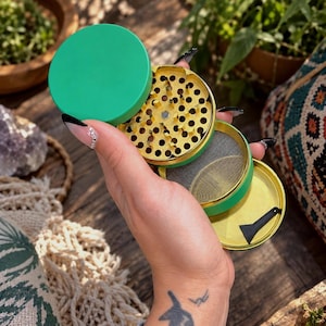 May include: A multi-piece herb grinder featuring a vibrant green exterior and a metallic gold interior. The device includes a top lid, a grinding section with sharp teeth, a fine mesh screen layer, and a bottom collection chamber with a small black scraper.
