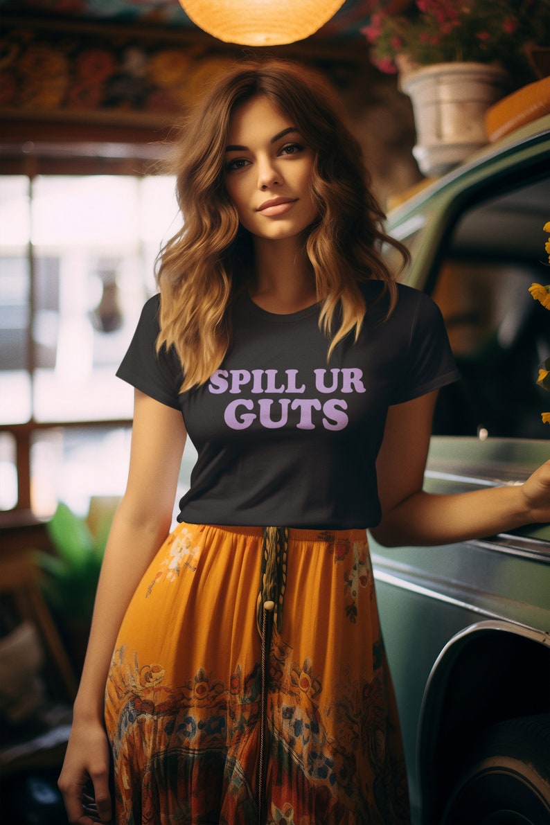 May include: A woman wearing a black t-shirt with the text "SPILL UR GUTS" in purple lettering. She is wearing a long orange skirt with a floral pattern and a brown belt.