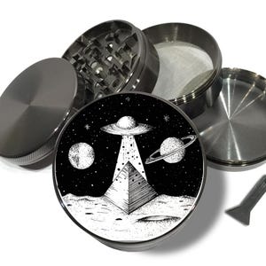 May include: A disassembled, silver-colored herb grinder with a black and white graphic of a UFO over a pyramid on the lid. The graphic also includes planets and stars. A small, silver scraper is included.