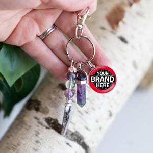 May include: A silver keychain with a crystal point, beads, and a clip. The keychain also has a round charm with the text "YOUR BRAND HERE" in black on a red background.