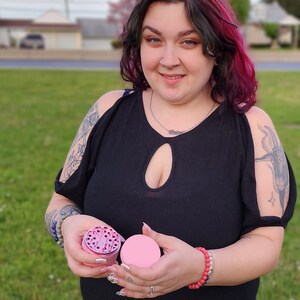 May include: A pink and silver grinder with a clear lid. The grinder is being held by a person with a tattoo on their arm.