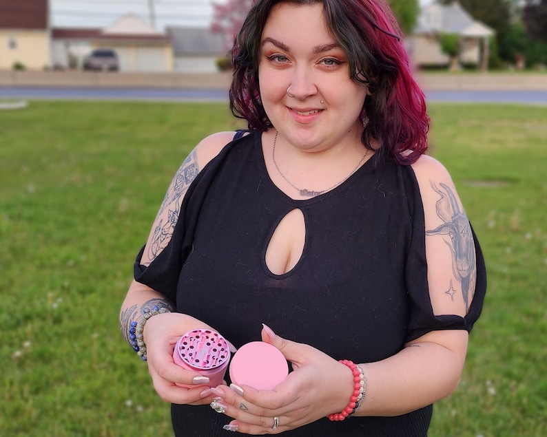 May include: A pink and silver grinder with a clear lid. The grinder is being held by a person with a tattoo on their arm.