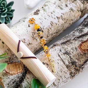 K&ouml;nnte beinhalten: Eine goldene Bienen-Haarklammer mit gelben und schwarzen Perlen, einem goldenen Blumenanh&auml;nger und einer goldenen Klammer. Die Klammer liegt auf einer Holzoberfl&auml;che.