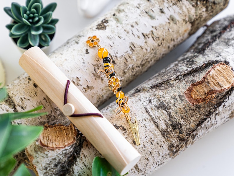 K&ouml;nnte beinhalten: Eine goldene Bienen-Haarklammer mit gelben und schwarzen Perlen, einem goldenen Blumenanh&auml;nger und einer goldenen Klammer. Die Klammer liegt auf einer Holzoberfl&auml;che.