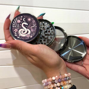 May include: A metal grinder with a floral design featuring a snake and moon phases. The grinder has a black and silver finish and comes with a screen and a scraper.