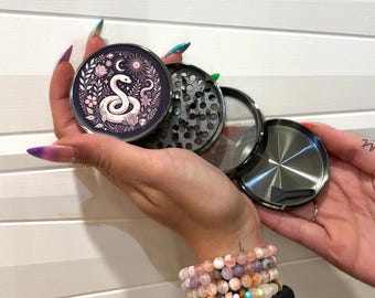 Puede incluir: Un molinillo de metal con un diseño floral que presenta una serpiente y fases lunares. El molinillo tiene un acabado negro y plateado y viene con una pantalla y un raspador.