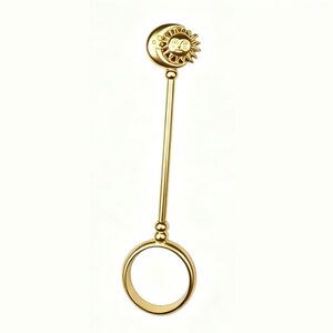 May include: A gold-colored ring holder with a celestial design. The top features a crescent moon and a sun with a smiling face. The ring holder has a long, straight stem and a circular ring at the bottom.