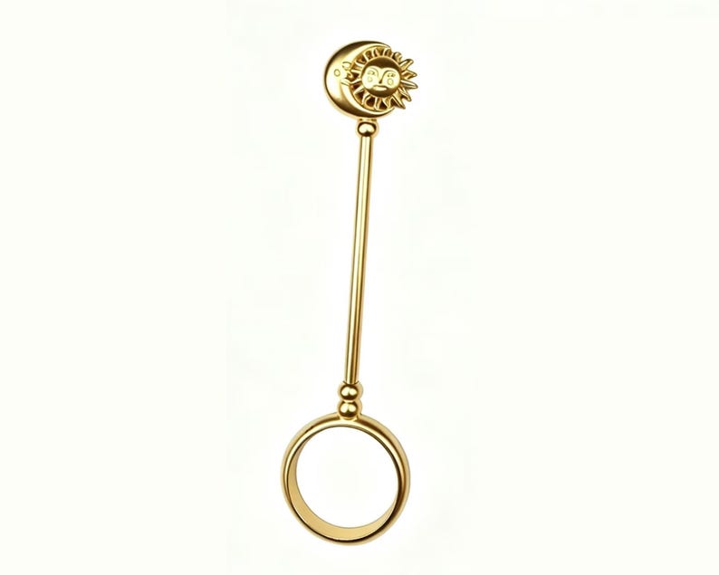 May include: A gold-colored ring holder with a celestial design. The top features a crescent moon and a sun with a smiling face. The ring holder has a long, straight stem and a circular ring at the bottom.