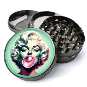May include: A metal grinder with a round, removable top featuring a colorful illustration of a woman blowing a bubble. The woman has blonde hair and is wearing a pink dress and earrings. The grinder has a silver finish and a green background.