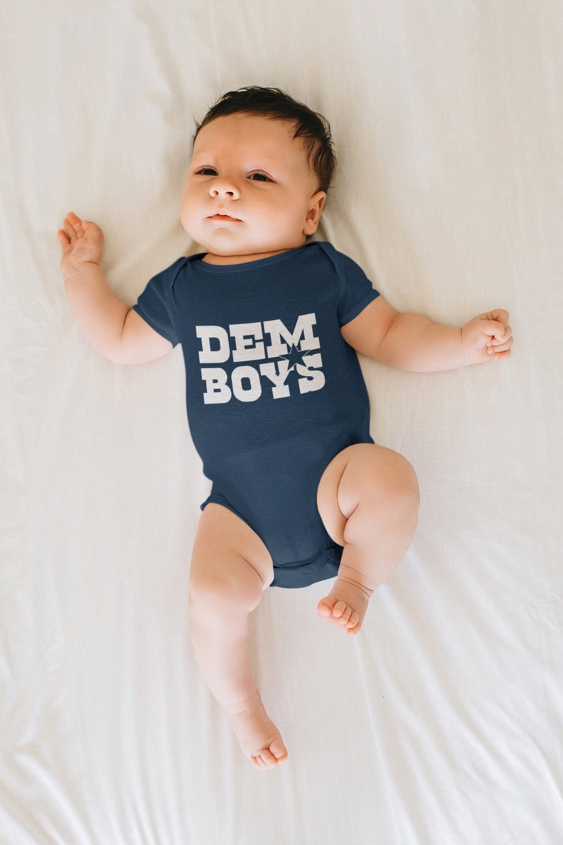 Pode incluir: Um beb&ecirc; vestindo um macac&atilde;o azul marinho com o texto "DEM BOYS" em letras brancas.