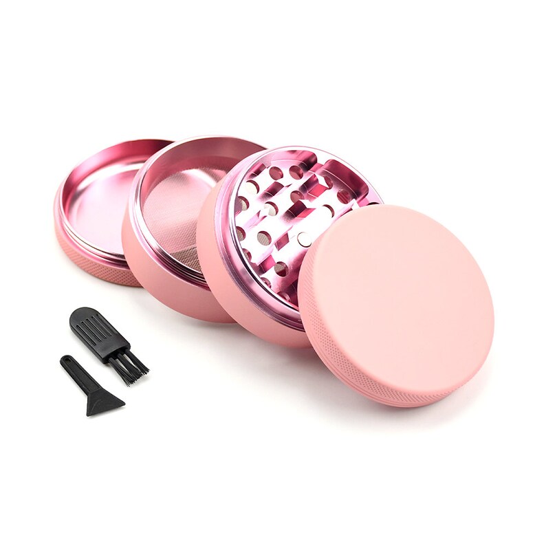 May include: A pink metal grinder with four pieces, including a top, a bottom, a grinder with teeth, and a screen. The grinder also includes a small black scraper and a brush.