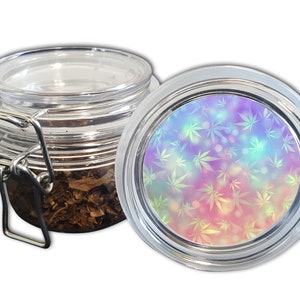Può includere: Un barattolo di vetro trasparente con coperchio incernierato, contenente foglie essiccate. Il coperchio presenta un design colorato di foglie di cannabis nelle tonalità del rosa, viola e verde. Il barattolo è progettato per la conservazione.