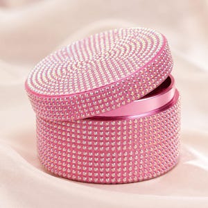 May include: A cylindrical pink metal grinder featuring a lid and base completely covered in small, reflective, light-catching rhinestones. The lid is slightly offset, revealing a smooth pink metallic interior rim within the circular container.