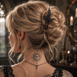 May include: A black metal hair clip shaped like a bird with a small red bead accent, worn in a blonde updo hairstyle. The accessory features intricate wing detailing and is positioned on the side of the hair, securing the bun in place.