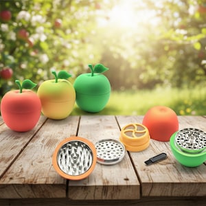 May include: A collection of apple-shaped grinders in red, yellow, and green, each with a green leaf top. The grinders are shown with their components, including a brush, on a wooden surface, with a blurred garden backdrop.