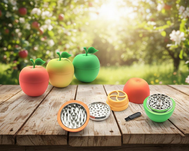 May include: A collection of apple-shaped grinders in red, yellow, and green, each with a green leaf top. The grinders are shown with their components, including a brush, on a wooden surface, with a blurred garden backdrop.