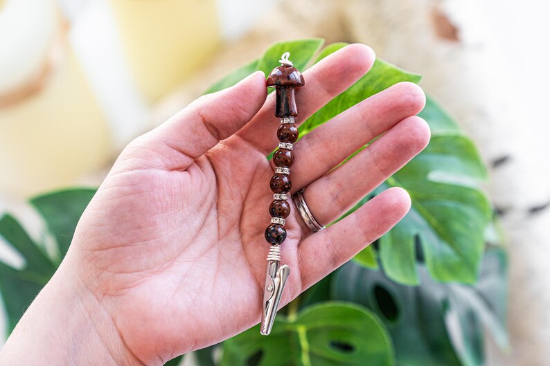 May include: A brown and silver beaded clip with a mushroom charm. The clip is made of metal and has a spring-loaded mechanism. The beads are brown and have a speckled pattern. The mushroom charm is brown and has a white stem.