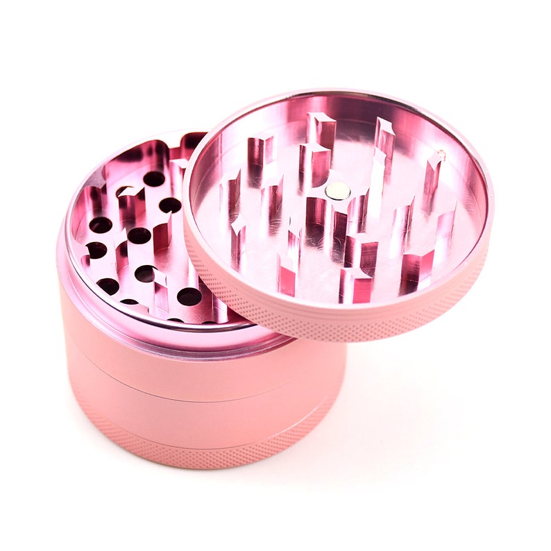 May include: A pink metal grinder with three sections. The top section is open and reveals the grinding teeth. The bottom section is closed and has a textured surface.