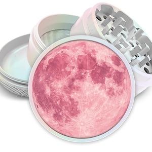May include: A white herb grinder with a pink moon design on the lid. The grinder is disassembled, showing the different components, including a mesh screen and grinding teeth. The moon graphic is a detailed illustration.