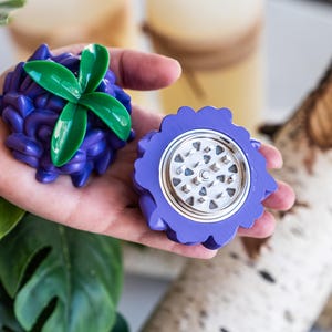 May include: Purple and green herb grinder shaped like a flower with a silver metal grinding mechanism.