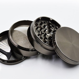 May include: A four-piece metal grinder with a black finish. The grinder has a top lid, a chamber with teeth, a screen, and a bottom chamber. The grinder also includes a small black scraper.