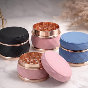 May include: Four colorful grinders with a geometric design. The grinders are in black, pink, and blue, with gold-colored accents. One pink grinder is open, revealing the grinding teeth.
