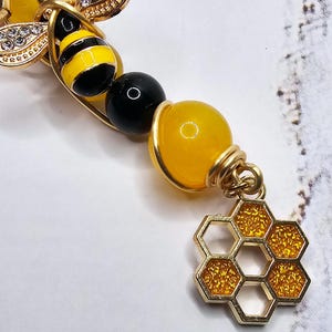 K&ouml;nnte beinhalten: Ein goldfarbenes Armband mit einem Bienen-Anh&auml;nger, einer Honigwabe und gelben und schwarzen Perlen.