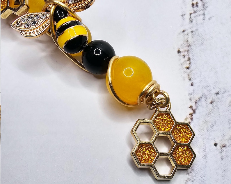 K&ouml;nnte beinhalten: Ein goldfarbenes Armband mit einem Bienen-Anh&auml;nger, einer Honigwabe und gelben und schwarzen Perlen.