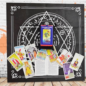 Może przedstawiać: Czarna szmatka do tarota z białym wzorem pentagramu i ozdobną obwódką. Niebieskie pudełko z napisem "Universal Waite Tarot Deck" leży na szmatce, otoczone kartami tarota z kolorowymi ilustracjami. Karty są rozłożone, prezentując różne obrazy.