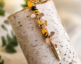 May include: A decorative roach clip featuring a gold-toned metal finish, adorned with yellow and black beads, two metallic bee charms with clear crystal accents, and two gold-toned honeycomb-shaped charms, ending in a functional metal alligator clip.