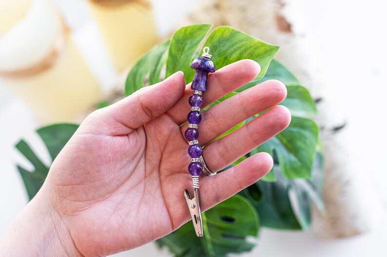 May include: A purple amethyst mushroom charm with a silver clip. The charm is made with amethyst beads and a silver wire.