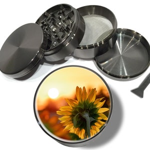 May include: A black metal grinder with a sunflower design on the bottom. The grinder has four parts: a lid, a top chamber, a bottom chamber, and a pollen catcher. It also includes a small metal scraper.