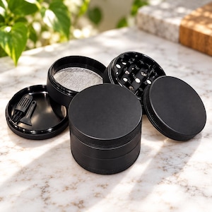 May include: A matte black four-piece herb grinder with a textured grip edge. The set includes a circular lid, a grinding chamber with sharp teeth, a fine mesh screen section, and a base compartment containing a small black cleaning brush and scraper.