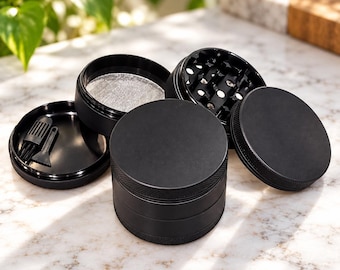 May include: A matte black four-piece herb grinder with a textured grip edge. The set includes a circular lid, a grinding chamber with sharp teeth, a fine mesh screen section, and a base compartment containing a small black cleaning brush and scraper.