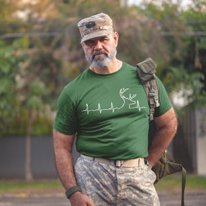 May include: A man wearing a green t-shirt with a white deer silhouette and a heartbeat graphic. He is also wearing a camouflage hat and pants.