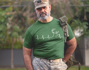 Pode incluir: Um homem vestindo uma camiseta verde com uma silhueta de veado branca e um gráfico de batimento cardíaco. Ele também está vestindo um chapéu e calças camuflados.