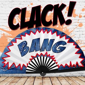 May include: A denim fan with a white and red burst design, featuring the word "BANG" in blue letters. The word "CLACK!" is written above the fan in black. The background includes a white brick wall with orange paint splatters.