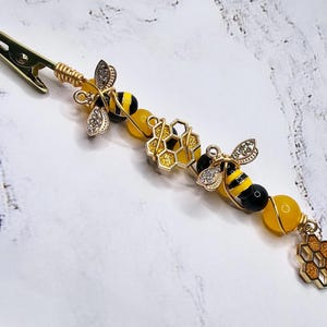K&ouml;nnte beinhalten: Eine goldene Bienen-Haarklammer mit schwarzen und gelben Bienen, goldenen Wabenanh&auml;ngern und gelben Perlen.