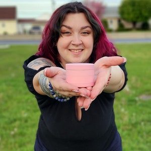 May include: A person is holding out a pink, three-tiered, round container. The container is made of plastic and has a smooth surface.