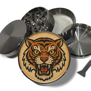 Peut inclure: Un broyeur métallique démonté avec un motif de visage de tigre détaillé sur le dessus. Le tigre a des rayures orange et noires, des yeux jaunes et une bouche rouge. Un petit grattoir noir est inclus.