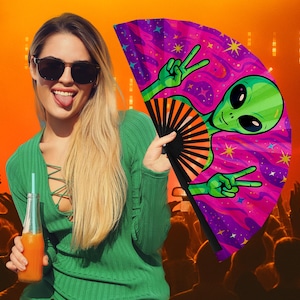 May include: A vibrant fan featuring a green alien with peace signs against a purple and pink cosmic background. The fan has a black handle and is held by a person wearing a green top and sunglasses, with a crowd in the background.