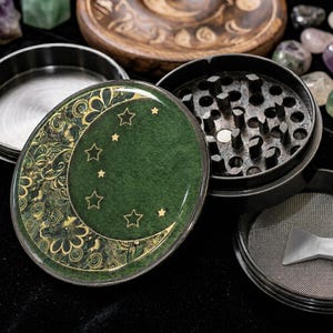 K&ouml;nnte beinhalten: Ein runder Kr&auml;uterm&uuml;hle aus Metall mit schwarzer Oberfl&auml;che. Der Deckel zeigt eine dunkelgr&uuml;ne Fl&auml;che mit einem goldenen Halbmond- und Sternenmuster, verziert mit komplexen floralen Details. Das Innere zeigt scharfe Z&auml;hne und ein Metallsieb.