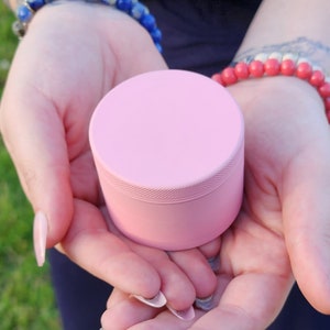 May include: A pink metal grinder with a smooth, rounded top and a textured bottom. The grinder is held in the hands of a person.