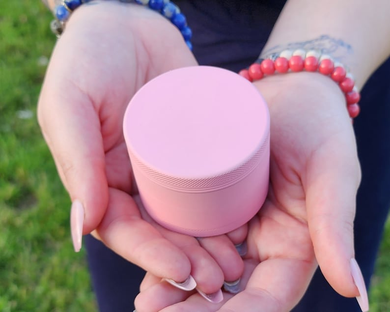 May include: A pink metal grinder with a smooth, rounded top and a textured bottom. The grinder is held in the hands of a person.