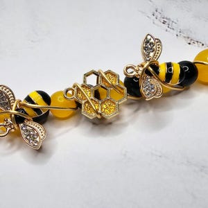 K&ouml;nnte beinhalten: Ein goldfarbenes, drahtgewickeltes Armband mit gelben und schwarzen Bienenanh&auml;ngern, einem goldenen Wabenanh&auml;nger und gelben und schwarzen Perlen.