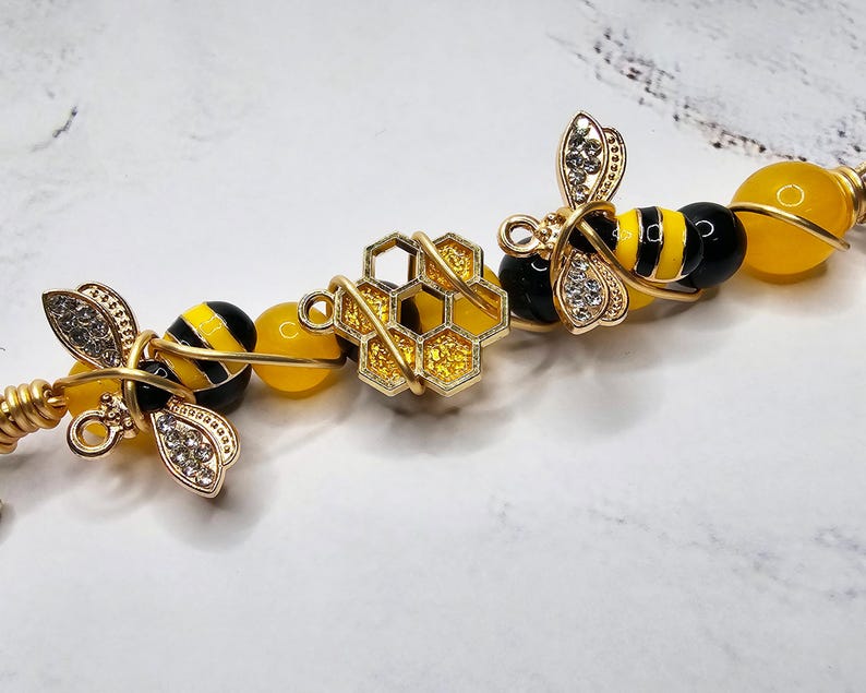 K&ouml;nnte beinhalten: Ein goldfarbenes, drahtgewickeltes Armband mit gelben und schwarzen Bienenanh&auml;ngern, einem goldenen Wabenanh&auml;nger und gelben und schwarzen Perlen.