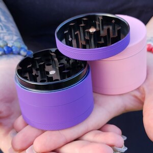 May include: Two metal grinders, one purple and one pink, with black metal grinders inside. The grinders are in the palm of a hand.