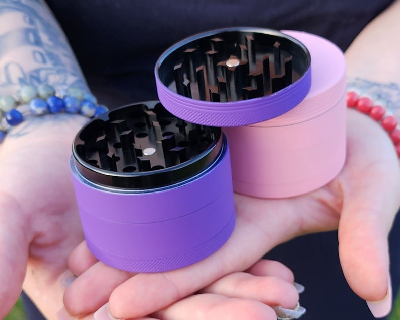 May include: Two metal grinders, one purple and one pink, with black metal grinders inside. The grinders are in the palm of a hand.